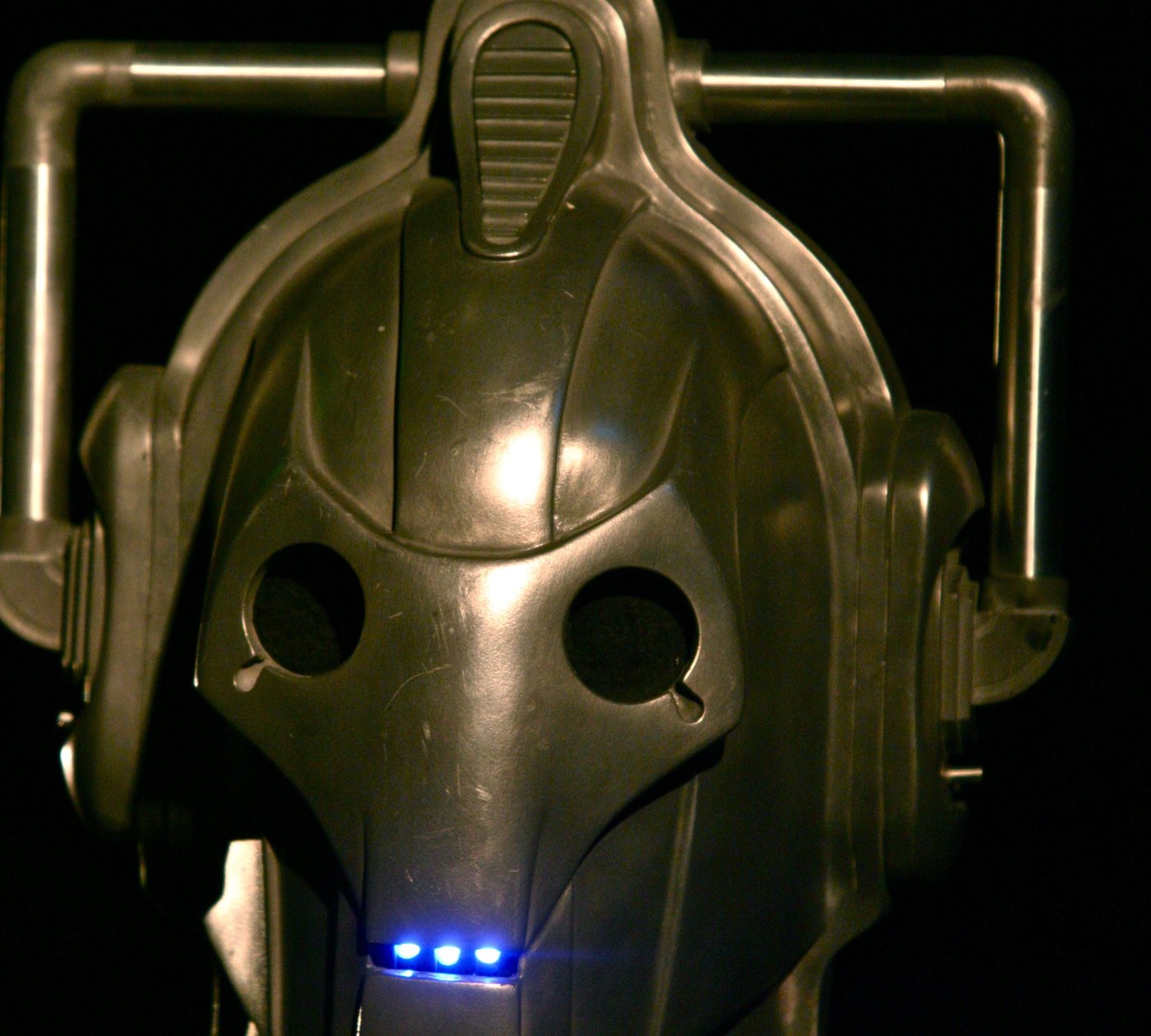 Close-up of a Cyberman helmet from Doctor Who, silver with blank round eyes and a blue light at the mouth