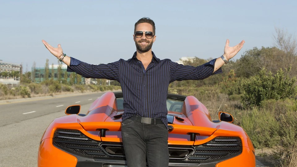 Russ Hanneman, played by Chris Diamantopoulos, standing arms outstretched in front of an orange McLaren in HBO's Silicon Valley